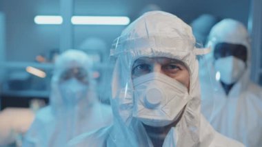 Beyaz tehlikeli madde giysileri, solunum cihazları ve yüz kalkanları içinde laboratuvarda kameraya bakan Kafkas doktor ve bulanık meslektaşlarının yakın plan portresi.