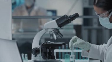 Modern laboratuarda mikroskop kullanarak koruyucu gözlük, eldiven ve maske takan genç kadın bilim adamının yan görüntüsü.
