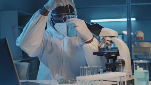 研究室の机に座っている白いハズマットスーツとマスクの男性黒科学者のウエストアップ 試験管内の有害化学物質を混合 — ストック動画