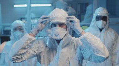 Laboratuvarda kameraya bakan beyaz koruyucu giysiler, solunum cihazları ve yüz kalkanları takmış üç farklı doktorun orta boy portresi.