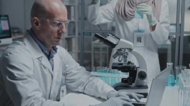 Yetişkin erkek bilim adamının orta ölçekli yakın çekimi, dizüstü bilgisayarda yazı yazması, kadın Müslüman asistanının test tüpünün yanında durup sıvıları karıştırması.