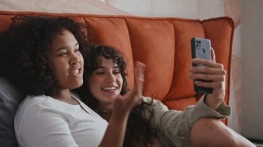 Modern etnik çeşitlilikteki lezbiyen çiftlerin yavaş çekimde aileleriyle veya arkadaşlarıyla akıllı telefon kullanarak evde oturup sohbet etmeleri.