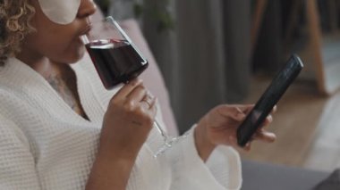 Göz altı yamalı modern Afro-Amerikalı kadın yatak odasında dinleniyor şarap içiyor ve akıllı telefondan sosyal ağda haber yayını yapıyor.
