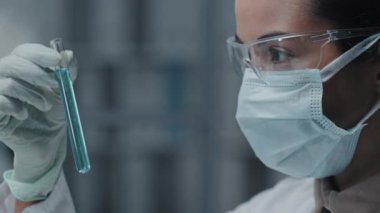 Eldiven, gözlük ve yüz maskesi takan kahverengi gözlü kadın bilim adamının modern laboratuarda sıvı içeren tüpünü yakın plan görüntüsü.