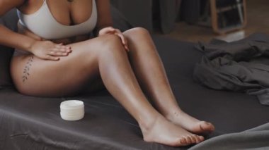 Genç siyahi bir kadının sabah yatak odasında bacaklarına nemlendirici krem sürerken orta boy fotoğrafı.