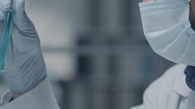 Eldiven, gözlük ve yüz maskesi takmış yetişkin bir bilim adamının mavi sıvıyla test tüpüne bakarken yan görüntüsü.