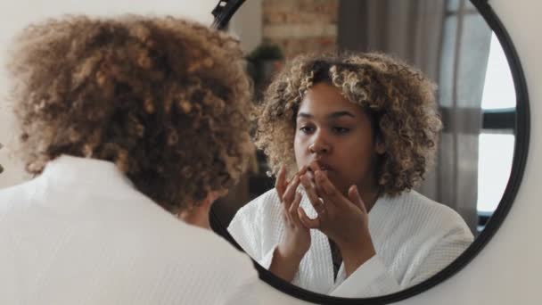 身穿白色长袍的年轻黑人妇女站在镜子前 涂上唇膏 触摸自己的面部肌肤 — 图库视频影像