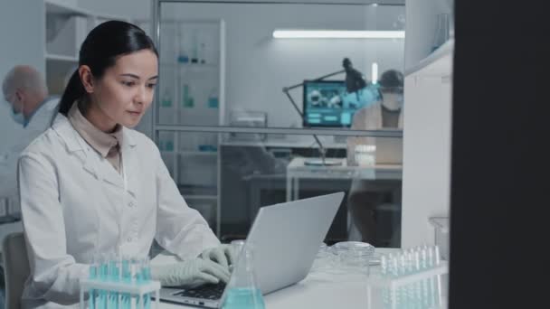 身穿白袍戴手套的年轻棕色眼睛女实验室技术员的蜡像 坐在办公桌前 手持计算机 看着相机 — 图库视频影像