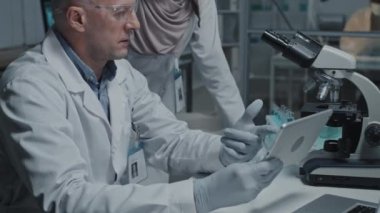 Yetişkin erkek bilim adamının masada oturup tablet bilgisayar kullanması, kadın Müslüman asistanının yanında durup konuşması.