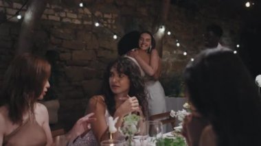 Gelinlik giymiş genç beyaz bir kadının, masada arkadaşlarıyla sohbet ederken, siyah karısının restoranda misafirlere sarılırken çekilmiş el resmi.