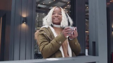 Sıradışı saç stili olan genç siyahi bir kadının portresi. Kışın verandada durmuş sıcak çay ya da kahve içiyor.