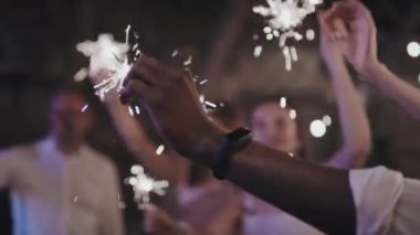 Seçici odak noktası, elinde maytap tutan etnik çeşitlilikteki insanların akşam partisinde dans etmesi.