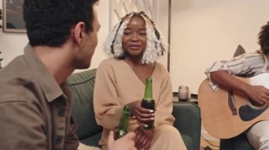 Oturma odasındaki kanepede oturan şık genç adam ve kadın ellerinde şişelerle parti sırasında arkadaşlarıyla sohbet ediyorlar.