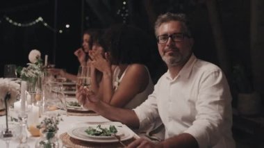 Beyaz gömlek ve gözlük takan yakışıklı, olgun beyaz bir adamın portresi düğün partisinde yemek masasında oturmuş kameraya bakıyor.