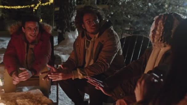 現代の民族的に多様な若い男性と女性キャンプファイヤーで手を暖め すべてについて話す屋外で霜の冬の夜を過ごす — ストック動画