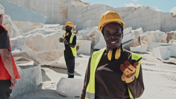 若い大人のアフリカ系アメリカ人女性のための選択的焦点中の肖像カメラを見てウォーキートーキーを保持する作業服 大理石の採石場の概念での作業日 — ストック動画