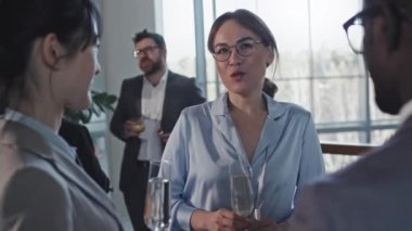 İş yerinde gözlük takan zarif bir kadın iş arkadaşlarıyla konuşurken elinde bir kadeh şampanya tutuyor.