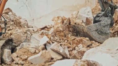 Tanınmayan kişi mermer ocağında kazıcı kullanıyor, kovayı taşlarla doldurmak ve kamyon yatağına taşımak için patlamayı kontrol ediyor.