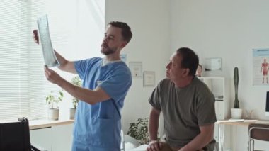 Üniformalı genç doktor röntgen görüntülerini gösteriyor ve hastaya hastaneye yaptığı ziyarette sorunu anlatıyor.