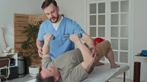 Fizjoterapeuta Mundurze Pokazuje Ćwiczenia Hantlami Dla Dojrzałego Pacjenta Leżącego Kanapie — Wideo stockowe