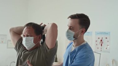 Maskeli yetişkin bir hasta rehabilitasyon sırasında halterle egzersiz yapıyor. Maskeli bir doktorla birlikte.