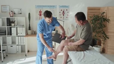 Üniformalı genç doktor, olgun bir hastanın yaralı bacağını inceliyor ve muayenede not alıyor.