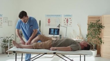 Genç fizyoterapist ofiste kanepede yatarken egzersiz yapıyor ve hasta bacaklarına masaj yapıyor.