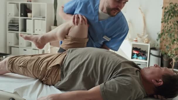 Młody Fizjoterapeuta Wykonujący Ćwiczenia Fizyczne Nogą Pacjenta Podczas Leżenia Kanapie — Wideo stockowe