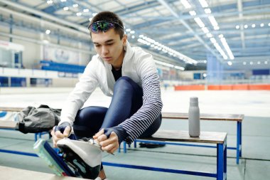 Aktif giyimli bir adam yedek kulübesinde oturuyor ve paten bağcıklarını bağlarken boş vakitlerinde buz arenasında yeni spor antrenmanlarına hazırlanıyor.