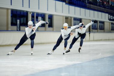 Spor üniformalı, patenli ve koruyucu gözlüklü üç genç sporcu buz pateni pistinde ilerlerken yarışmaya katılıyorlar.