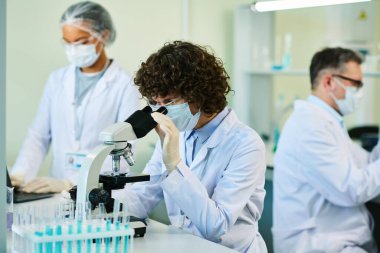 Laboratuvar önlüğü ve maskeli olgun esmer kadın, iş arkadaşları arasında yeni virüsün hücrelerini incelerken mikroskopla bakıyor.