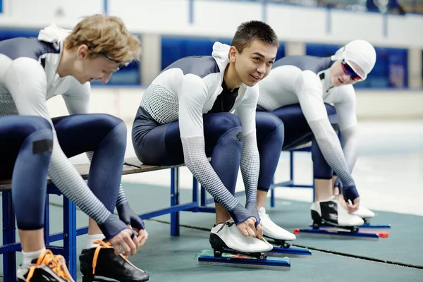 三个身穿运动服 身穿运动服的人 在休息的时候把鞋带系在冰鞋上 坐在长椅上排成一排 准备下一场比赛 — 图库照片