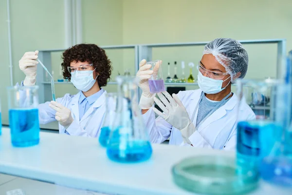 研究室のコートと保護マスクの2人の異文化科学者は 研究室で液体化学物質の特性を研究する — ストック写真