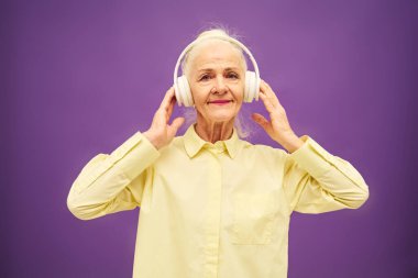Sarı tişörtlü mutlu bir kadın kulaklıkla müzik dinliyor ve menekşe arkaplanda poz verirken gülümseyerek kameraya bakıyor.