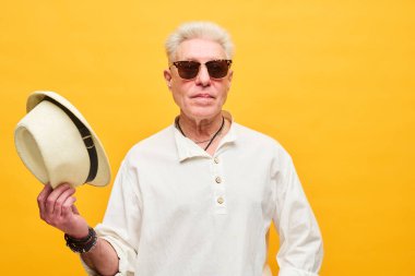 Güneş gözlüklü, beyaz gömlekli, sağ elinde panama şapkası olan, sarı arka planda kameranın önünde duran şık bir son sınıf öğrencisi.