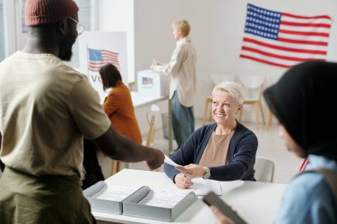 Seçim komisyonunun masasında duran Afrikalı Amerikalı genç adama oy pusulası kağıdı veren mutlu sarışın kadın.