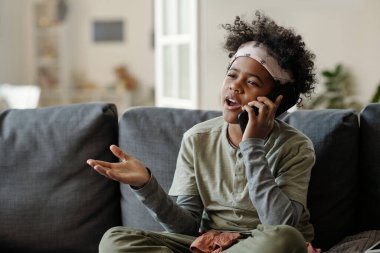 Akıllı telefonlu tatlı çocuk oturma odasında kanepede oturuyor ve cep telefonuyla konuşurken bir şeyler anlatıyor.