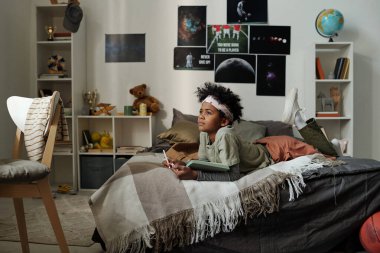 Ev kıyafetlerinden ilham alan okul çocuğu çift kişilik yatakta yatar ve okul ödevi için yeni fikirler düşünür ya da diğer ev ödevlerini yaparken