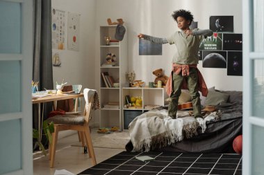 Afro-Amerikan okul çocuğu çift kişilik yatakta duvarlara yaslanmış posterler asıyor ve evde spor yaparken egzersiz yapıyor.