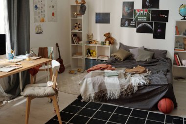 Çift kişilik yataklı geniş yatak odasının bir kısmı duvarın yanında duruyor. Posterler, raflar, oyuncaklar, kitaplar, malzemeler ve sandalyeyle dolu bir masa.