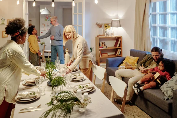 在为周末的家庭晚宴做准备的过程中 成熟和年轻的跨文化女性将盘子和酒杯放在客厅的桌子上 — 图库照片