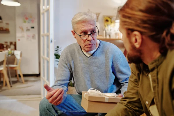 在客厅里 一个头戴礼品盒的成熟男人坐在儿子面前的沙发上 一边和儿子说话 一边解释着谈话中的一些事情 — 图库照片