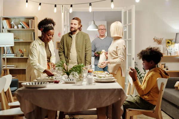当男人的父母从厨房端酒的时候 文化间的年轻丈夫和妻子在客厅里为家庭晚餐供应餐桌 — 图库照片