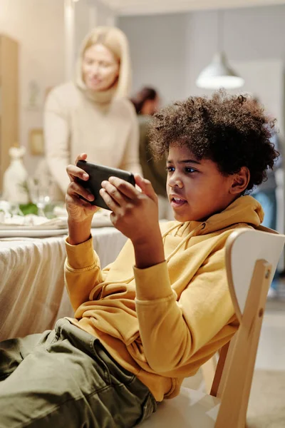 可爱的非洲裔美国男孩坐在椅子上与祖母一起吃饭 一边用智能手机观看网上视频 — 图库照片
