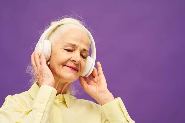 白い髪をした笑顔のシニア女性がヘッドフォンに触れながらラベンダーを背景に立ち 彼女の好きな音楽を楽しむ — ストック写真