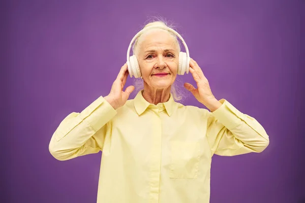 穿着黄色衬衫 快乐的老妇人 一边听着耳机里的音乐 一边微笑着看着相机 一边在紫色的背景上摆出一副姿势 — 图库照片