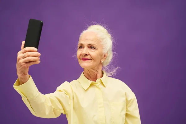 Старшая Улыбающаяся Женщина Жёлтой Рубашке Смотрит Экран Смартфона Делая Селфи — стоковое фото
