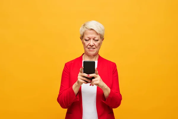 头发短小的金发妇女一边看智能手机屏幕 一边在黄色背景的视频聊天中发短信或交流 — 图库照片