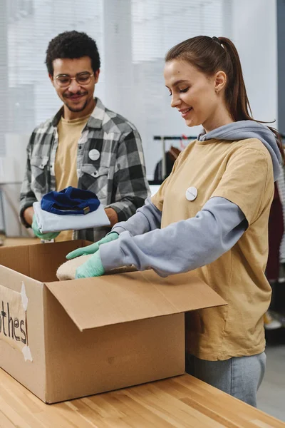 ボランティア団体の事務所の大きな箱に詰め込むために必要な人のための無料の服を準備する若い異文化ボランティア — ストック写真
