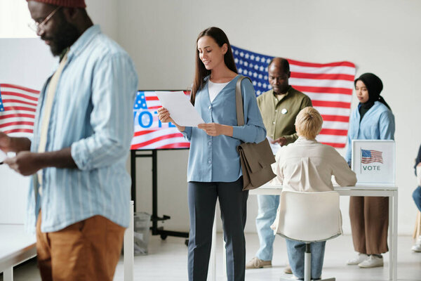 Happy young woman looking through ballot paper while standing in queue at polling place among intercultural citizens of USA